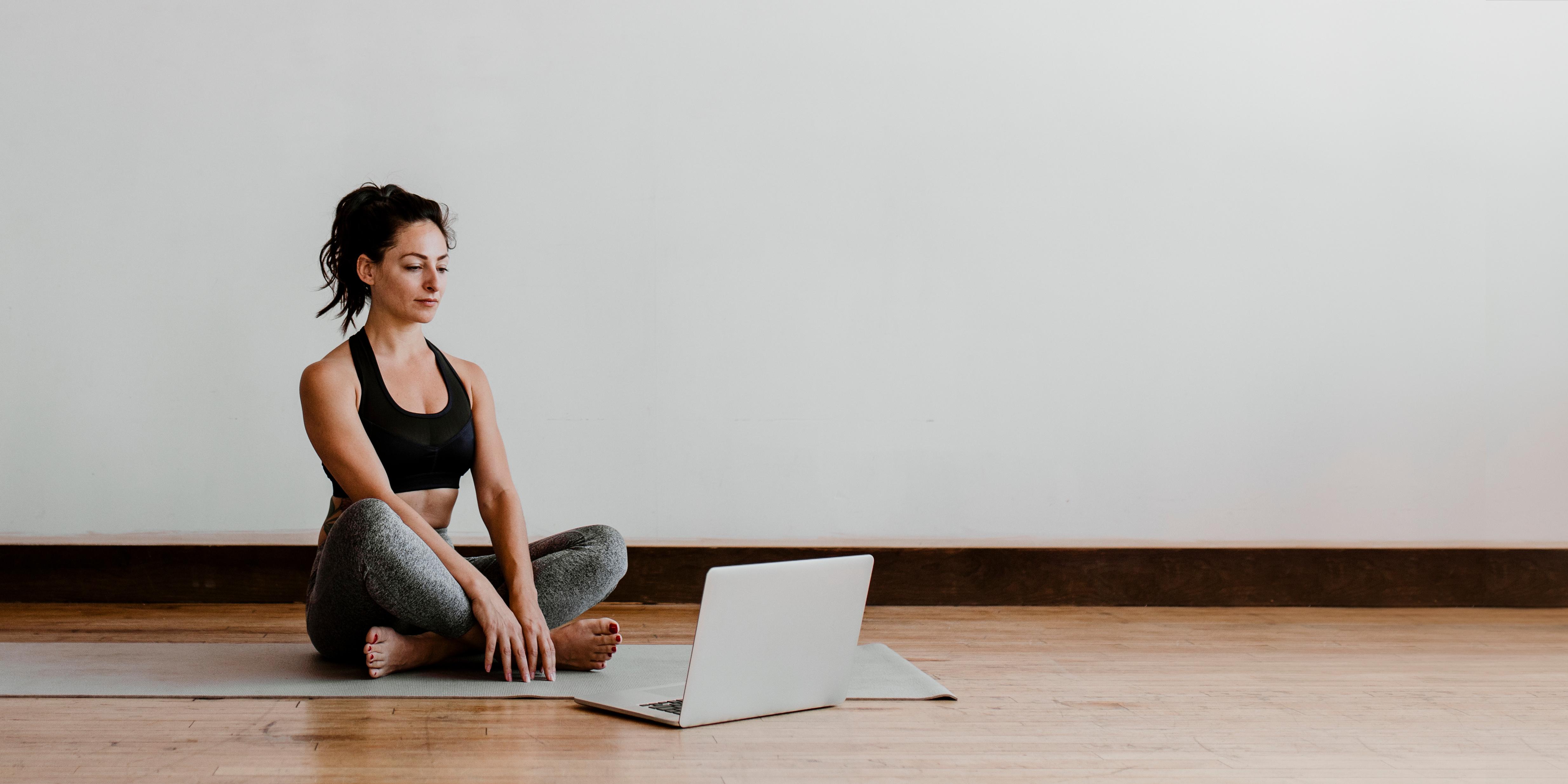 An adult sits in a yoga posture in front of a laptop