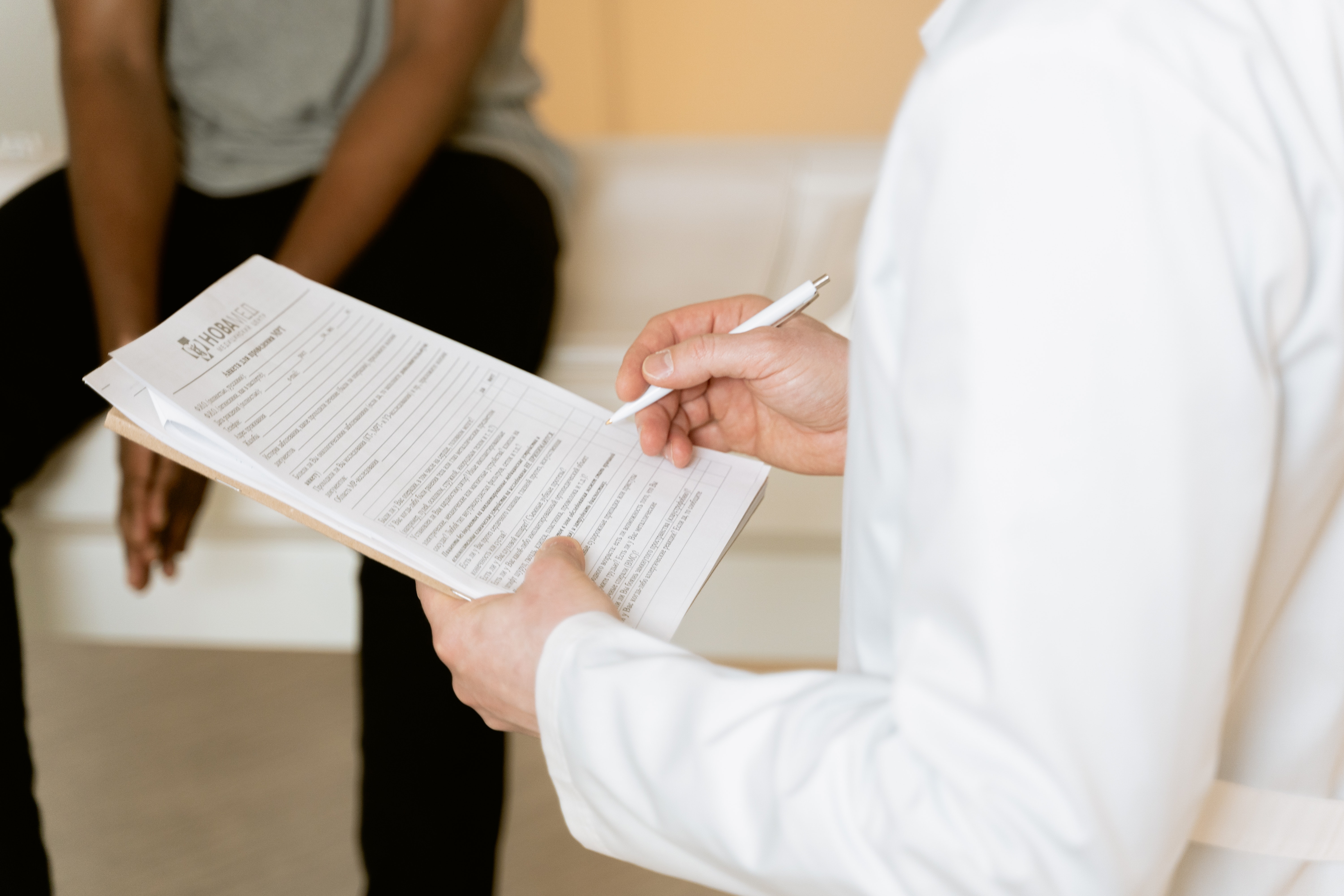 A person in a medical coat holds a clipboard and pen. A person in the background is seated.